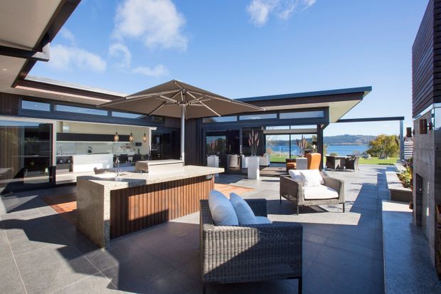 A contemporary outdoor entertaining terrace centered on a built-in outdoor kitchen island with a wood-slatted base and stone benchtop. The paved courtyard includes woven lounge chairs, an umbrella, planters, and covered architectural pavilions with large glazed openings. A body of water and distant hills are visible beyond the property.