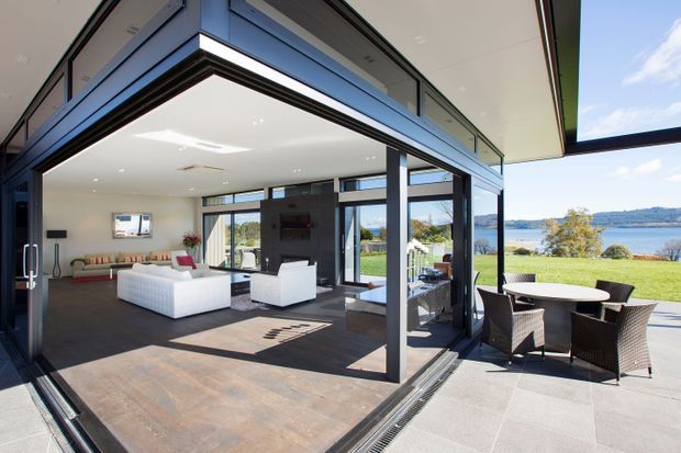 A bright contemporary indoor-outdoor living space with large retractable glazed openings connecting a furnished living room to a covered terrace. The interior has dark flooring, a white sectional sofa, white armchairs, a fireplace or media wall, and expansive glazing. Outside, a dining table and woven chairs overlook a landscaped lawn and a body of water. Clean-lined detailing, slim dark frames, and a broad overhanging roof create a strong connection to the landscape.