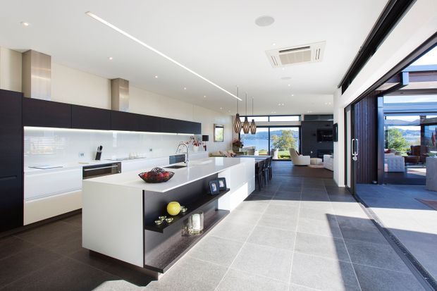 A bright, open-plan contemporary kitchen with a large white island, dark timber cabinetry, integrated appliances and a long illuminated splashback. The space extends into a dining and living area with pendant lighting, while expansive black-framed sliding glass doors open to an outdoor terrace. Large-format grey floor tiles and abundant natural light create a clean, spacious interior.