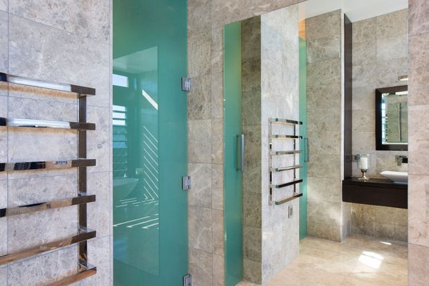 A contemporary bathroom clad extensively in large-format beige and grey natural-stone-look tiles. The room features a frameless turquoise-tinted glass shower enclosure, multiple wall-mounted heated towel rails, a floating dark-wood vanity with a white basin, and a large wall mirror. The space is bright, polished, and spa-like, with strong vertical divisions and reflective surfaces.