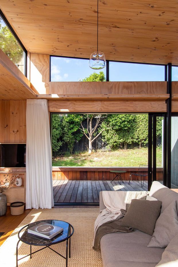 Warm, compact living room with extensive timber lining on the ceiling and walls, black-framed glazing, and a large sliding glass door opening onto a timber deck and leafy garden. A neutral sofa, woven rug, round coffee table, and pendant light create a relaxed contemporary interior. High-level clerestory windows and a partial loft-like opening bring in abundant natural light.