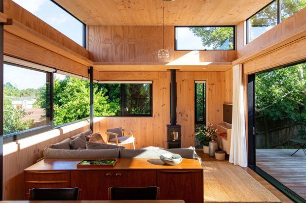 A warm, timber-lined open-plan living and dining space with extensive black-framed glazing, a compact wood-burning stove, built-in timber shelving, and mid-century-style furniture. The room opens directly onto a timber deck and leafy garden, with a raised ceiling and clerestory-like upper windows providing abundant natural light.