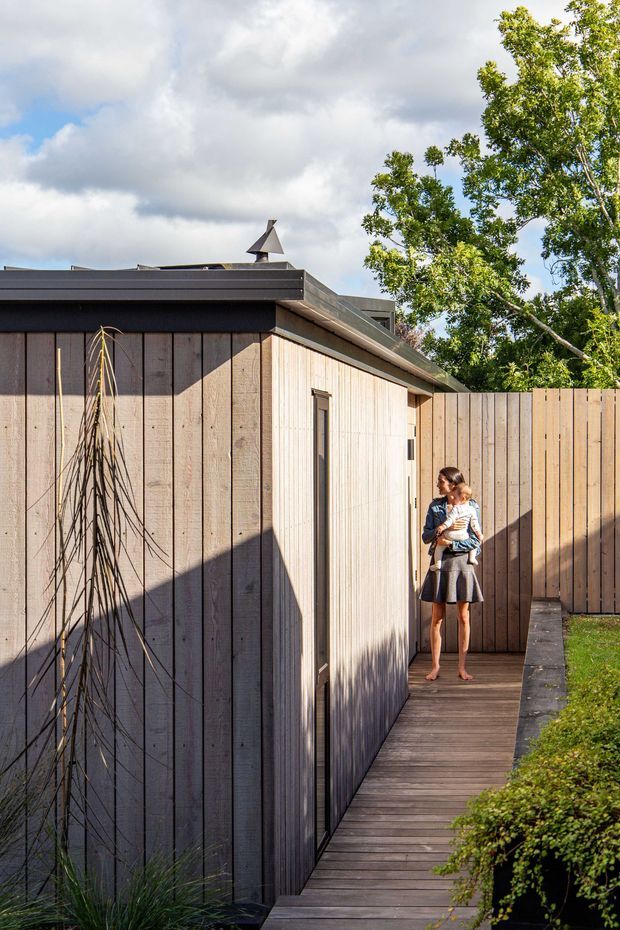 Exterior view of a compact contemporary building with dark vertical timber cladding, a lighter timber-clad projecting volume, and a low-profile dark metal roof. A narrow timber boardwalk runs alongside the structure and meets a matching timber privacy fence, with landscaped planting at the edges. A person is present for scale. Bright natural daylight and strong shadows emphasize the textured facade and clean geometric form.