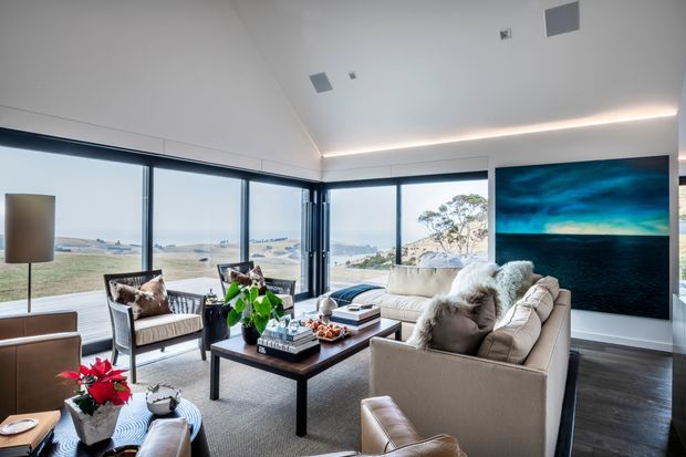 A refined, light-filled living room with floor-to-ceiling corner glazing overlooking an open coastal or rural landscape. The room features multiple cream upholstered sofas and armchairs, a dark rectangular coffee table, dark timber flooring, a large abstract blue artwork, and concealed linear ceiling lighting. The neutral furnishings and expansive glazing create an airy, contemporary entertaining space.