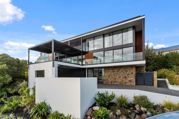 Contemporary multi-level residence with a dramatic sloping roofline, extensive floor-to-ceiling glazing, dark metal framing, timber wall accents, and a prominent natural stone masonry wall. The home includes a covered outdoor terrace or carport-like structure, layered white rendered walls, landscaped planting, and a glimpse of a swimming pool in the foreground.