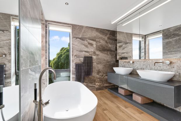 Luxurious contemporary bathroom with a freestanding white bathtub, double wall-mounted vanity with vessel basins, large mirror, brushed metal fixtures, dark stone-look wall tiling, small mosaic backsplash, and warm timber-look flooring. A window brings in natural light and greenery views.
