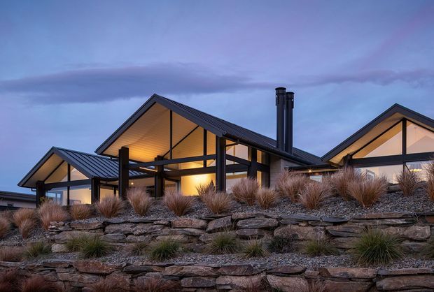 Twilight exterior view of a contemporary, single-storey gabled house set behind layered natural-stone retaining walls and ornamental grasses. The home features dark standing-seam metal roofs, extensive warm-lit glazing, exposed dark structural framing, and multiple intersecting pitched roof forms.