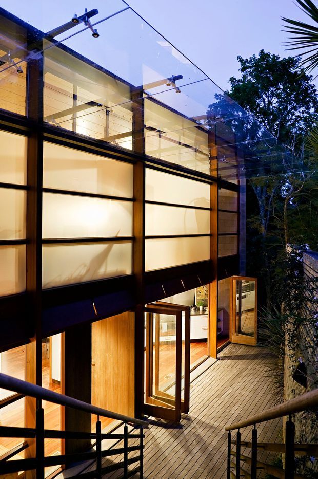 A dramatic view of a multi-level modern building entrance and terrace at dusk. Large translucent illuminated wall panels are framed by dark structural members, with extensive glazing, exposed timber ceilings, and warm timber flooring and doors. A glass canopy with exposed mounting hardware shelters the upper level, while stairs and timber decking lead to open glazed doors. Dense vegetation is visible beyond the building.