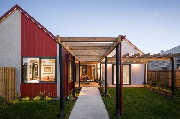 Exterior view of a contemporary residential property with a red vertical-clad gabled volume, pale rendered walls, timber pergola-covered walkway, concrete path, lawn, and timber fencing. Glazed doors and windows reveal a warm interior dining or living area, while a second gabled volume and low roof forms extend behind the courtyard-like approach.