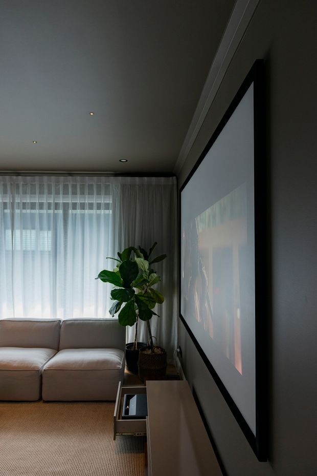 A dimly lit contemporary living room featuring a large wall-mounted projection screen or television, a low linear media console, a pale sectional sofa, sheer full-height curtains, recessed ceiling lights, and a large indoor leafy plant. The room has a calm, intimate home-cinema feel with neutral finishes and warm timber-toned flooring.