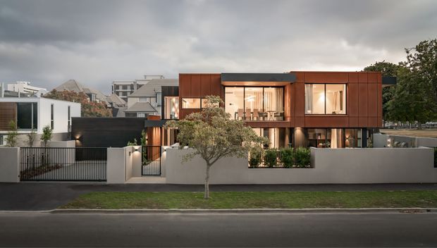Street-facing view of a contemporary multi-level residence with a strong rectilinear composition, dark metal roof elements, warm rust-brown cladding, expansive glazed openings, and a pale rendered boundary wall. The home includes a recessed upper-level terrace or balcony, landscaped planting, a gated driveway, and a small front garden. Overcast skies and warm interior lighting create a refined evening architectural presentation.