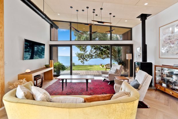 A bright, double-height living room with a large curved sectional sofa, armchairs, coffee table, media console, and red area rug. Floor-to-ceiling glazing opens to a timber deck and lawn with a visible coastal water view. The room features exposed black services and a freestanding black fireplace flue, with contemporary pendant lighting and warm timber flooring.