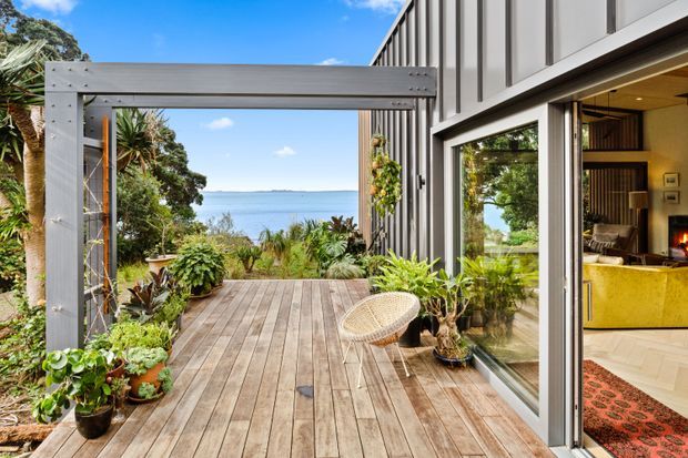 A contemporary coastal home exterior with a timber deck opening toward a calm body of water. The deck is framed by a grey timber pergola or portal structure, abundant potted greenery, and vertical dark cladding. Large glazed sliding doors reveal a warmly styled living room with timber cabinetry and a fireplace inside.