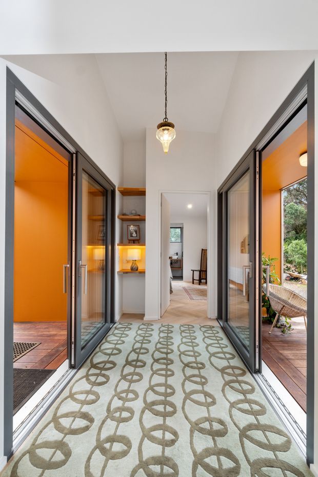 A bright transitional hallway or entry passage with full-height glazed folding doors opening onto timber-floored spaces on both sides. The corridor has white walls and ceiling, dark metal door frames, a patterned runner, recessed display shelving with warm lighting, and a hanging pendant light. An additional room is visible at the far end.