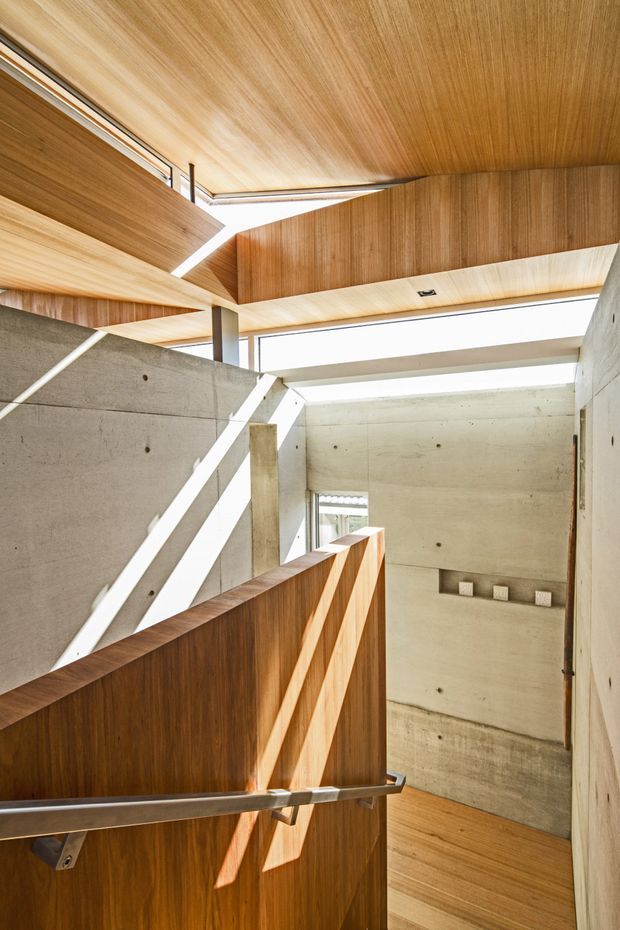 Interior architectural photograph of a stairway or landing with a warm timber-clad ceiling, timber balustrade, exposed smooth concrete walls, narrow clerestory glazing, and strong angular sunlight. The composition emphasizes the sculptural junction of timber, concrete, daylight, and stairs, with a clean contemporary material palette.