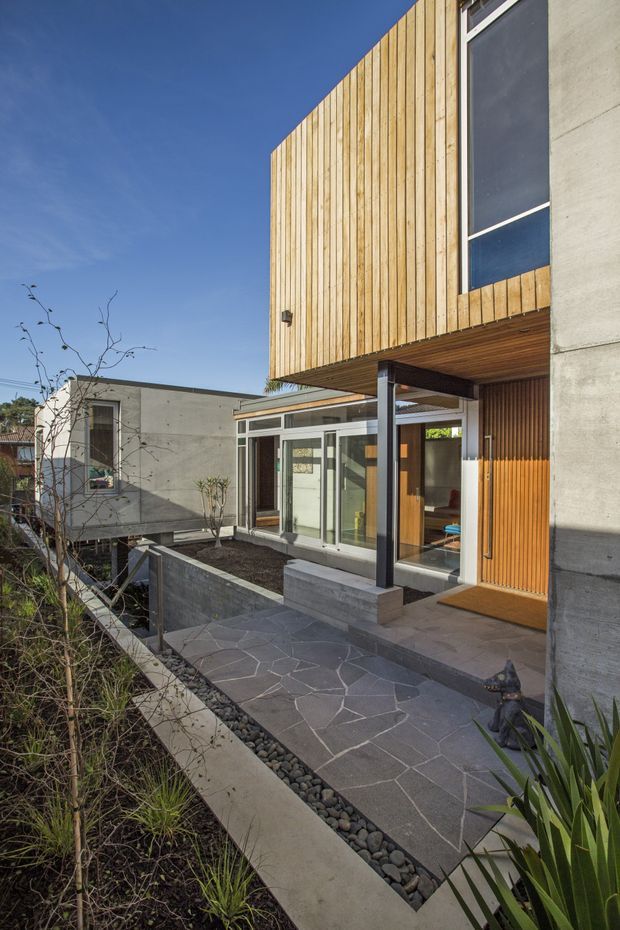 Exterior architectural photograph of a contemporary multi-level residence viewed from a landscaped approach. The house combines vertical timber cladding, exposed smooth concrete walls, large glazed doors and windows, and a projecting upper volume. A paved stone entry terrace and walkway are bordered by dark pebbles and structured planting, with an internal courtyard-like arrangement visible beyond the entrance.