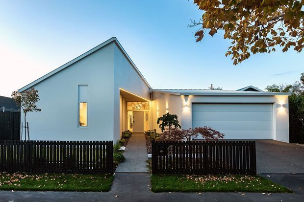 Contemporary single-storey suburban home photographed from the street at dusk. The pale rendered gable-fronted dwelling has a recessed, warmly lit entry walkway and an attached double garage with a broad sectional door. Dark vertical timber fencing, a concrete driveway and path, clipped planting, and a small front lawn frame the facade.