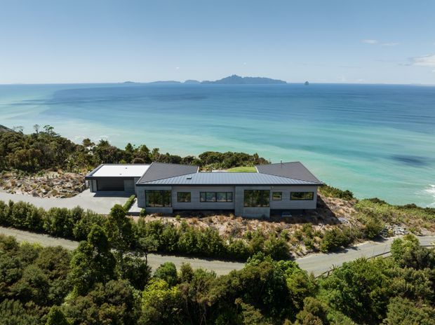 Aerial view of a low-profile contemporary coastal house set among native vegetation on a rocky shoreline, with expansive turquoise ocean views and a distant island or headland beyond. The home has a simple linear form, dark pitched roofs, grey masonry or cladding, and extensive horizontal glazing.