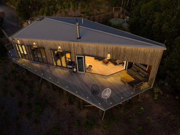 Aerial dusk view of a contemporary elevated timber-clad house set among dense bush. The building has a broad low-pitched roof, extensive glazing, a large wraparound timber deck, outdoor seating, and warm interior and exterior lighting. A chimney and outdoor grill are visible on the deck.
