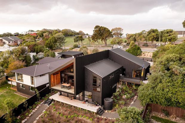 Aerial view of a contemporary multi-volume residential house set within a leafy suburban landscape. The home combines dark vertical cladding and dark metal roofing with a lighter rendered or weatherboard-like volume, warm timber around the recessed entry and balcony, and extensive garden planting. Several pitched and skillion roof forms, decks, and large glazed openings are visible.