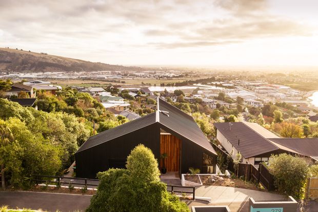 Aerial exterior view of a contemporary dark-clad house on a sloping suburban site, with a steep gabled roof, prominent timber entry door, and landscaped surroundings. The elevated viewpoint overlooks a dense residential neighbourhood, broad waterway, and hills in warm late-day light.