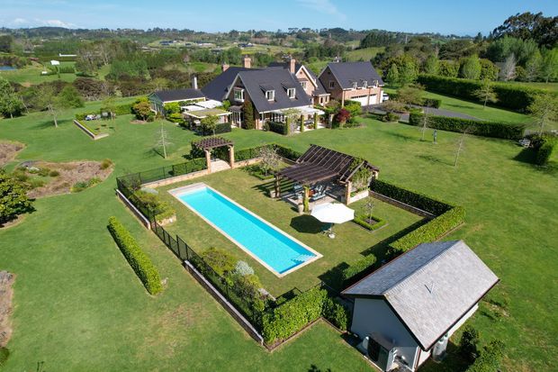 Aerial view of a large rural residential property with a dark pitched-roof house, expansive manicured lawns, hedges, landscaped gardens, a rectangular swimming pool, pergola and detached outbuilding. The surrounding countryside and neighboring fields are visible beyond the property.