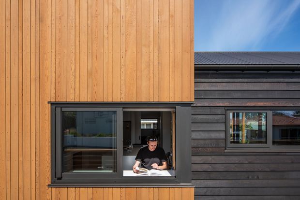 Contemporary building facade clad in warm vertical timber boards and dark horizontal weatherboards. A projecting or recessed window opening forms a small study or reading nook, with a person seated at a desk inside. The composition highlights contrasting timber cladding textures, dark window frames, and a low-pitched roof edge against a blue sky.