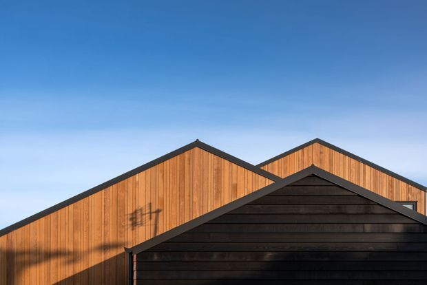 Close-up exterior view of overlapping gabled building forms with vertical warm timber cladding and a dark horizontal-clad foreground gable. Strong geometric rooflines are set against a clear blue sky.