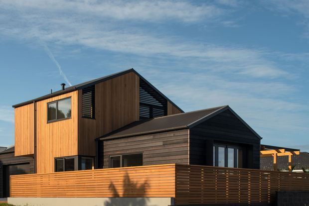 Exterior view of a contemporary multi-level house with a steep gabled roof, contrasting vertical timber cladding on the upper volume and dark horizontal cladding on the lower front volume. A horizontal timber privacy fence and dark-framed windows define the foreground facade.