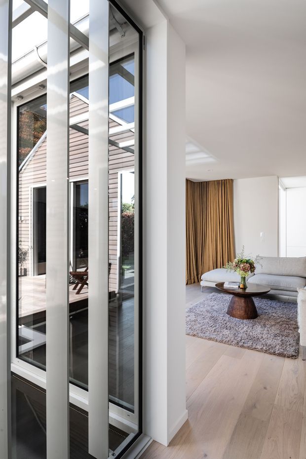 A bright contemporary living room viewed beside a dramatic floor-to-ceiling glazed atrium or entry zone. White structural columns and slim dark window frames create strong vertical lines, while a glazed roof brings in abundant daylight. The living room contains a pale grey sofa, textured grey rug, round dark timber coffee table, and full-height warm tan curtains. Light timber flooring continues through the space. Through the glazing, a pale timber-clad exterior courtyard or adjoining structure is visible.