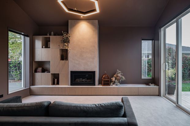 A dark, sophisticated living room with a full-width built-in timber media and fireplace wall, a textured central fireplace surround, low integrated bench cabinetry, and a black sectional sofa. Large windows and sliding glass doors bring in views of the garden, while a geometric LED ceiling pendant provides a contemporary focal point.