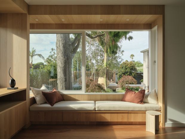 A warm, contemporary built-in window seat framed by extensive timber joinery. The upholstered bench has neutral cushions and rust-coloured accent pillows, with a large picture window overlooking a leafy garden. Integrated shelving and cabinetry occupy the left side, while recessed ceiling spotlights and timber-lined reveals create a refined, cohesive interior.