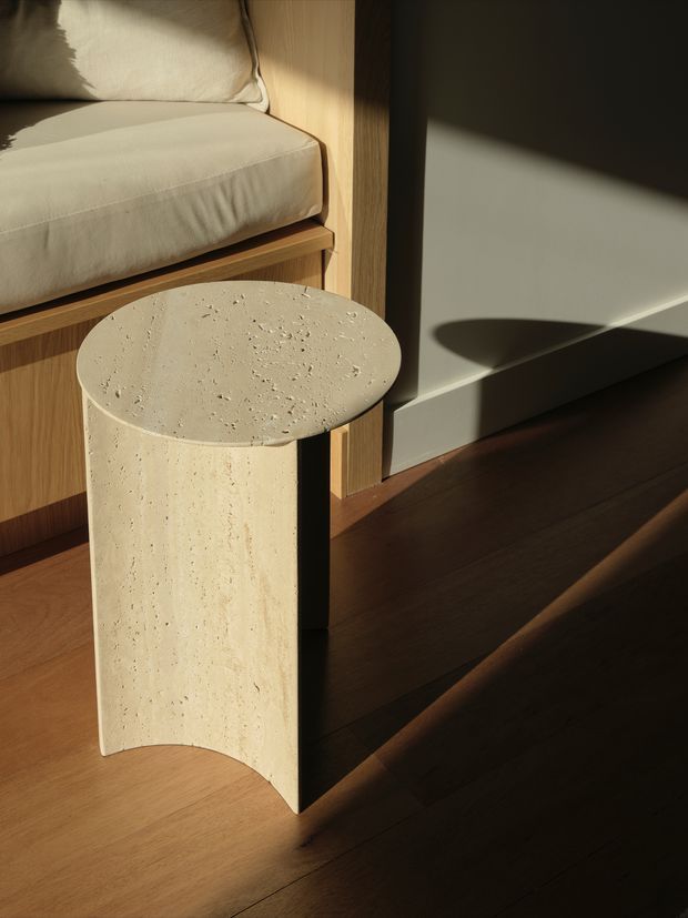 Close-up interior detail of a small cylindrical side table with a heavily textured, light cream stone or travertine-like surface, positioned beside a built-in wooden bench with cream upholstery. Warm sunlight casts strong shadows across the dark timber flooring and pale wall, creating a refined, minimalist composition.