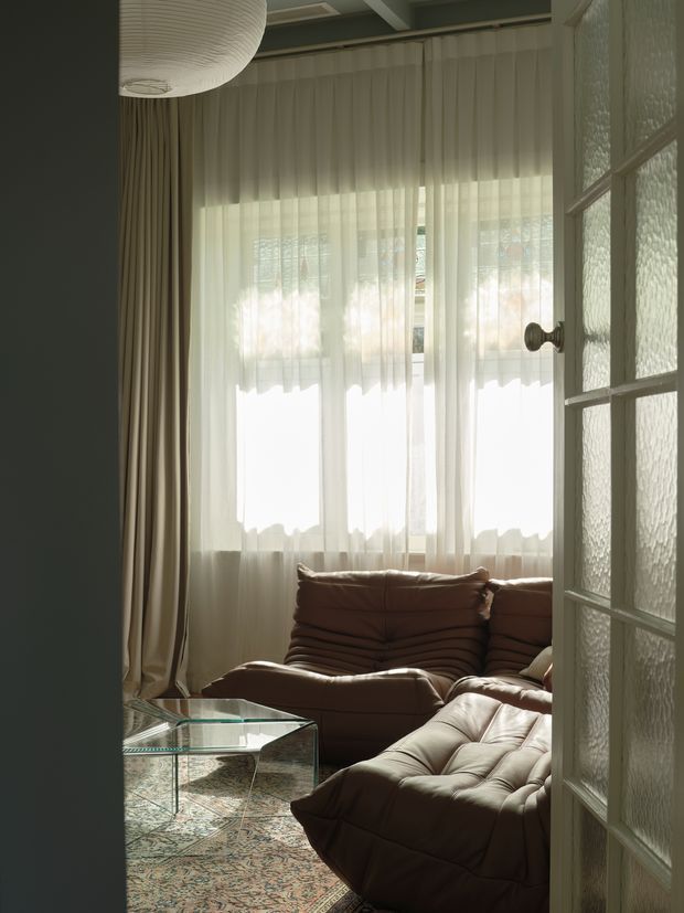 A warmly lit living room viewed through a partially open multi-pane textured-glass door. The room features deep brown, channel-tufted leather seating, a transparent glass coffee table, full-height sheer curtains with heavy side drapes, and a large sculptural pendant light. The space has a cozy, softly layered vintage-inspired atmosphere.