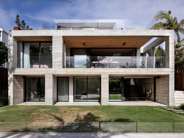 Front exterior view of a contemporary two-storey concrete residence with a strong rectangular frame, broad glazing, recessed balconies, glass balustrades, timber-lined soffits, and a rooftop terrace. A landscaped lawn and palm vegetation sit in front of the house.