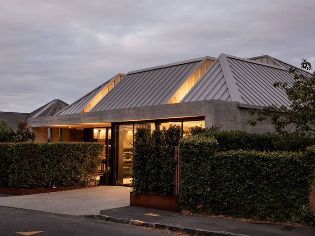 Twilight exterior view of a low-rise residential building with a complex series of steeply pitched metal gable roofs, clerestory-like illuminated roof openings, and a broad concrete or masonry entry canopy. Warm interior lighting glows through the glazed entrance, partially screened by clipped hedges and a paved forecourt along the street.