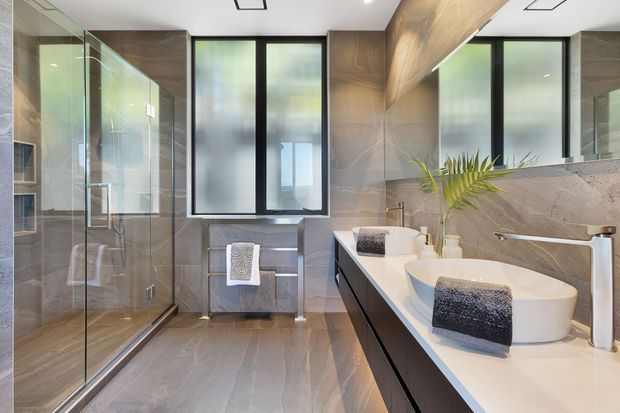 Contemporary bathroom with a spacious frameless glass shower enclosure, freestanding white bathtub, double vanity, and wall-to-wall grey stone-look surfaces. Black-framed frosted windows, brushed metal fixtures, recessed lighting, and dark vanity cabinetry create a refined, modern fit-out.