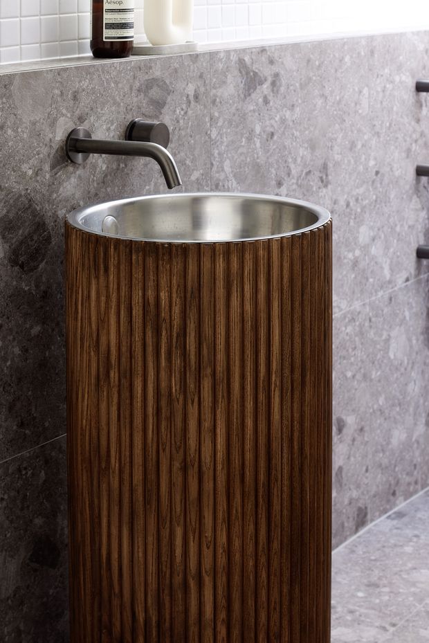 Close-up of a refined bathroom vanity featuring a tall cylindrical fluted dark timber cabinet with an inset brushed stainless-steel basin. A wall-mounted dark metal tap projects over the basin, while large grey stone-look wall and floor tiles create a cohesive backdrop. A narrow ledge with toiletries sits beneath a white square-tile upper wall.