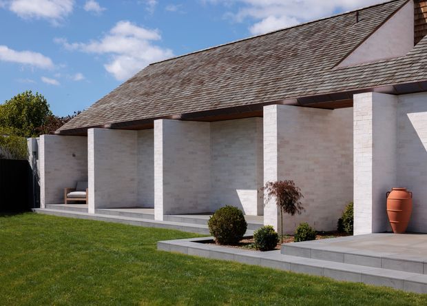 Exterior view of a low, modern pavilion-like building with a broad steeply pitched shingle roof, pale masonry walls, deep covered verandah, and regularly spaced square columns. A paved terrace, clipped shrubs, lawn, and a large terracotta vessel frame the facade under a bright blue sky.