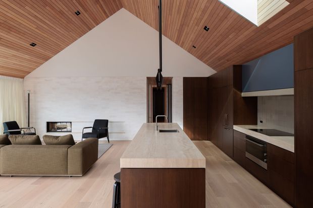 Open-plan contemporary kitchen and living area with a large central island, dark timber cabinetry, pale timber flooring, and a dramatic vaulted timber-lined ceiling. The kitchen includes integrated appliances, a stone benchtop, and a dark geometric rangehood. A compact living zone with a sofa, armchairs, and a linear fireplace is visible beyond the island.