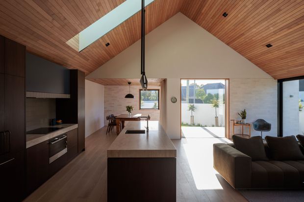 Open-plan contemporary living, dining and kitchen interior with a dramatic timber-lined pitched ceiling and long skylight. A dark built-in kitchen occupies the left side, with a central island in a pale stone-like finish, dining table beyond, and a large dark sofa in the foreground. Full-height glazing and a large window bring in strong natural light, with a small courtyard visible outside.