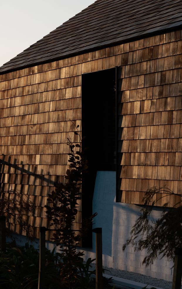 Close-up exterior view of a compact building clad in overlapping timber shingles, with a steeply pitched shingle roof and a tall, dark recessed doorway. A pale painted masonry wall is visible beneath the cladding, with planting and strong angled shadows in the foreground. The image has a dramatic, warm, low-light architectural photography style.