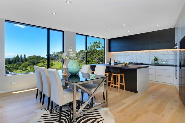 Bright open-plan dining and kitchen interior with floor-to-ceiling windows overlooking a leafy landscape. The space features pale timber flooring, a sleek black kitchen island and cabinetry, white lower cabinets, a dark tiled or panelled splashback with warm under-cabinet lighting, a glass dining table with upholstered white chairs, and contemporary recessed ceiling lights.
