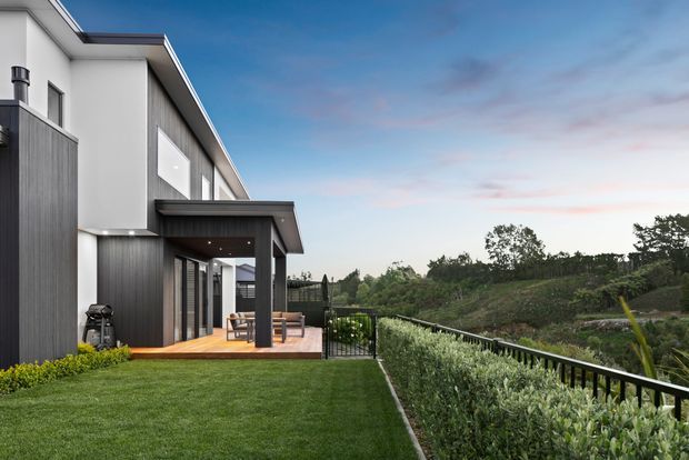 Contemporary multi-level residential house viewed from the landscaped rear yard, with a crisp white rendered upper volume, dark vertical cladding, broad roof overhang, covered outdoor terrace, timber decking, black metal balustrade, and expansive green outlook. The image is photographed at dusk with a calm suburban or semi-rural setting.