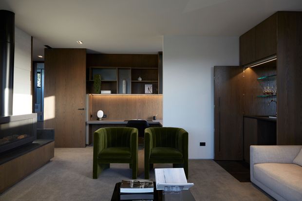 A refined contemporary living room with a dark timber entertainment wall and built-in shelving, a concealed bar or kitchenette cabinet at right, two deep green upholstered armchairs, a pale sofa, and a low glass coffee table. Soft integrated lighting and warm timber joinery create a calm, moody interior.