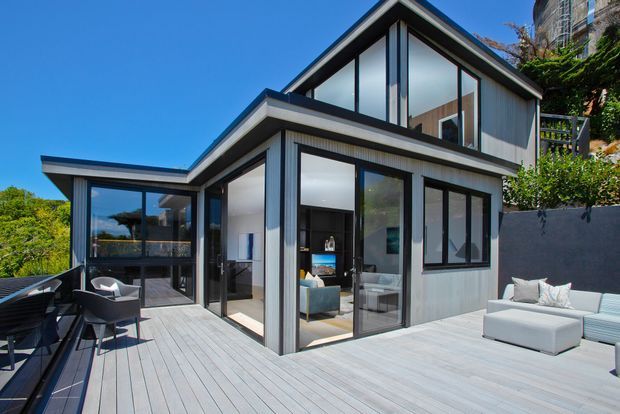 Contemporary multi-level residential house viewed from a large outdoor timber deck. The home features strong angular roof forms, extensive black-framed floor-to-ceiling glazing, light vertical cladding, and open-plan interiors visible through the windows. Outdoor dining and lounge furniture occupy the spacious deck, with landscaped terrain and neighbouring structures in the background.