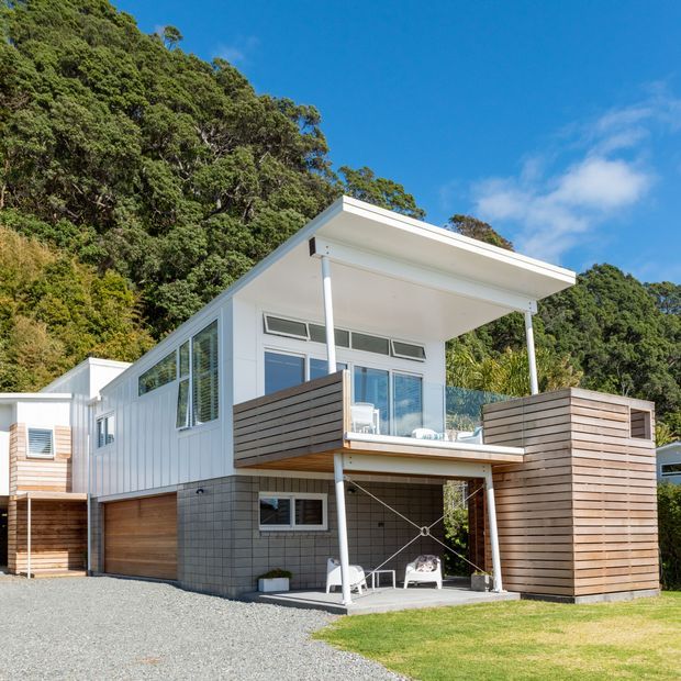 A modern multi-level house set against a densely wooded hillside. The white-clad structure has a broad flat roof, extensive glazing, a raised upper living level, timber privacy screens and balconies, a ground-level garage, and an open sheltered outdoor area beneath the upper floor.