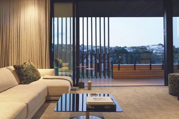 A refined living room with a large cream sectional sofa, textured vertical timber wall panelling, a low reflective coffee table, and floor-to-ceiling glazing opening to a balcony or terrace. Dark slim window frames and vertical exterior screening frame views across the surrounding landscape and buildings. The room has a warm, contemporary feel with soft neutral furnishings and integrated architectural detailing.
