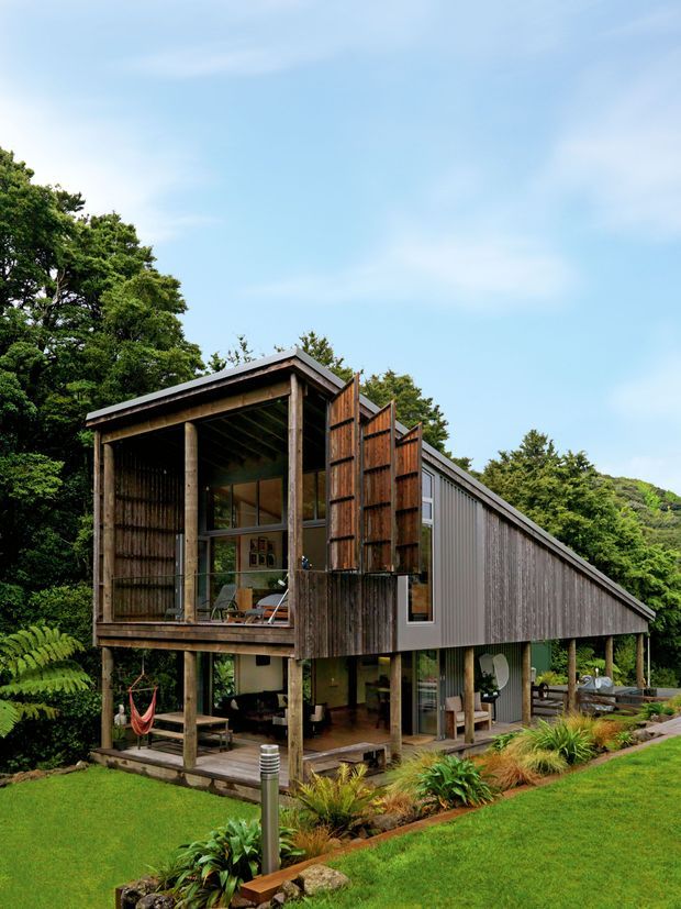 A contemporary elevated house set within a lush, wooded landscape. The building features a long sloping roof, extensive weathered timber screening and exposed timber posts, with an open-sided lower level and a glazed upper living area. Native-style planting, ferns and lawn surround the structure.