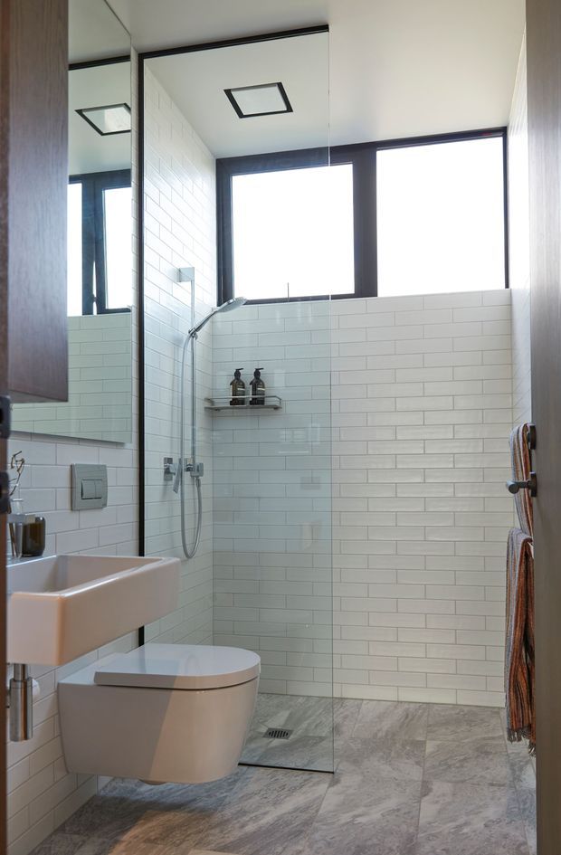 A compact contemporary bathroom featuring a frameless walk-in shower with a clear glass screen, white subway-tiled walls, a wall-hung toilet, and a wall-mounted basin. Large black-framed windows provide natural light, while grey stone-look floor tiles and a mirrored wall create a clean, functional interior.